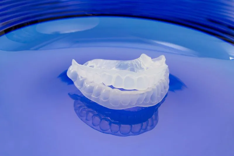 Claro, custom-molded dental night guard for the upper teeth resting on a reflective blue surface.